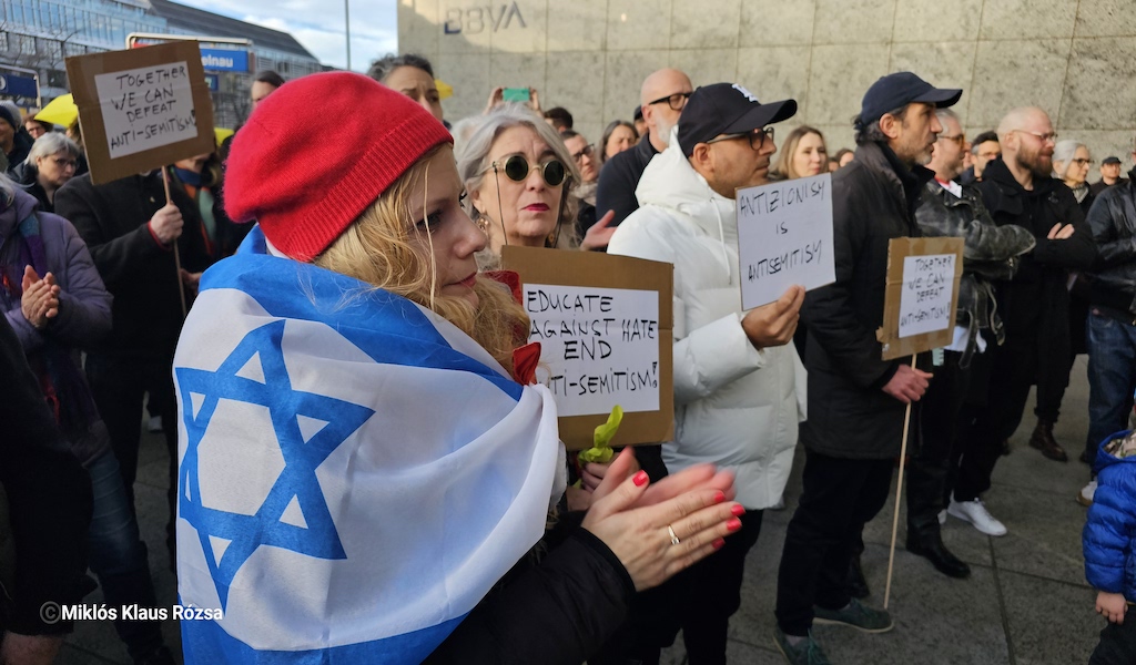 Eine Frau trägt eine israelische Flagge um die Schultern und klatscht bei einer Kundgebung, während andere Teilnehmende Schilder gegen Antisemitismus hochhalten.