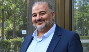 A portrait of Mansour Abbas wearing a dark blue blazer and a light blue shirt, standing in front of a glass facade with green trees in the background.